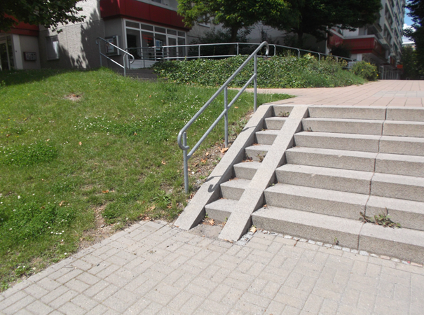Das Foto zeigt eine steile Rampe, die am Rand auf einer Treppe angelegt wurde. Die Treppe wird seitlich, neben der Rampe durch ein nicht kontrastierendes Geländer begrenzt. Die Rampe besteht, für die, rechten und linken Räderpaare eines Rollstuhls, aus jeweils ca. 10 cm breiten schiefen Ebenen, die sich vom Boden vor der untersten Stufe bis zur Trittfläche der obersten Stufe erstrecken. Der Zwischenraum zwischen beiden schiefen Ebenen ist mit Stufen versehen.