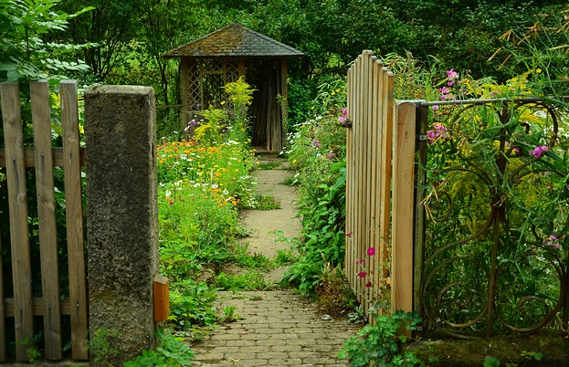 Bildbeschreibung: Das Foto gewährt den Blick in einen Bauerngarten. Das Gartentor aus Holz auf der rechten Bildseite ist geöffnet. In der Bildmitte führt ein Plattenweg, der zum Teil mit Pflanzen bewachsen ist, auf einen Gartenpavillon aus Holz mit einem Spitzdach zu. Rechts und links des Weges wachsen verschiedene Blumen in unterschiedlicher Höhe und bunten Blütenfarben, wie rosa und gelb. Ende der Bildbeschreibung.