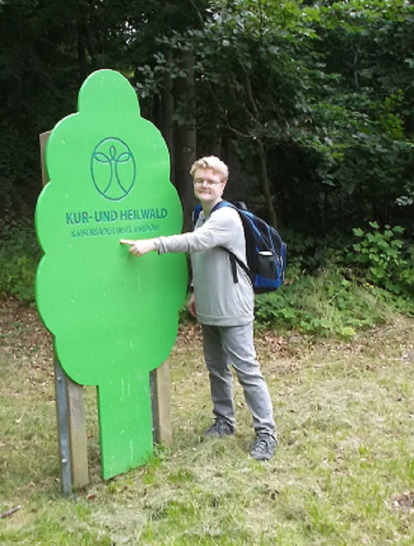 Bildbeschreibung: Auf Bild 3 zeigt Claudia auf den Schriftzug Kur - und Heilwald der sich auf dem hellgrünen Eingangsschild in Form eines Laubbaumes befindet. Dieses Schild steht am Eingang von Europas 1. Kur- und Heilwald in Heringsdorf auf Usedom. Ende der Bildbeschreibung.