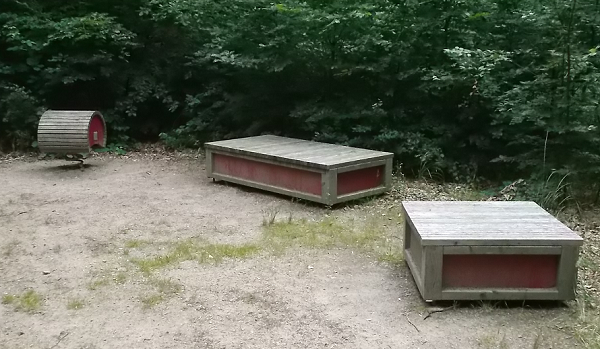 Bildbeschreibung: Auf Bild 7 sind zwei quaderförmige und eine runde Sitzgelegenheit, je aus mehreren Holzbrettern bestehend, zu sehen, unter denen sich eine Feder befindet. Damit sind die Sitzgelegenheiten im Waldboden verankert. Ende der Bildbeschreibung