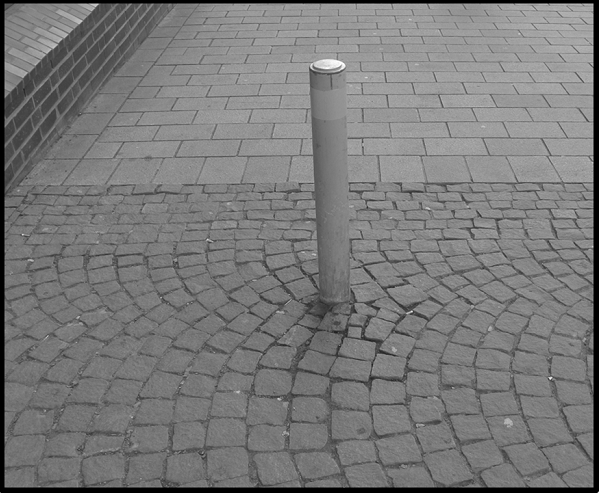 Bildbeschreibung: Bild 2 zeigt in der Mitte eines, mit grauen Granitsteinen, gepflasterten Gehweges einen grauen Poller / Sperrpfosten. Ende der Beschreibung.