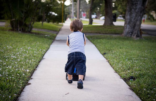 Bildbeschreibung: Bild 1 zeigt einen kleinen Jungen von etwa 2 Jahren, der auf einem Gehweg vom Betrachter wegläuft. Der Betonweg ist recht und links von Rasen umgeben, auf dem Bäume und Sträucher zu sehen. Ende der Beschreibung. 