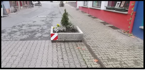 Bildbeschreibung: Das Bild 10 zeigt einen rechteckigen Pflanzkübel aus Beton in der Mitte eines verkehrsberuhigten Bereichs stehen. An seiner linken Seite ist er mit einem rot-weiß-schraffierten Rechteck gekennzeichnet. Ende der Bildbeschreibung. 