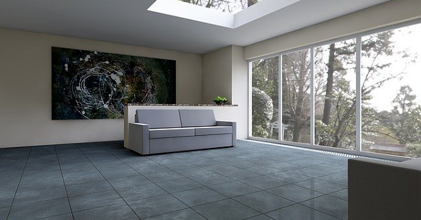 Bildbeschreibung: Das Bild zeigt einen Wohnraum mit einem Dachfenster und einer Glasfassade rechts. Auf den grauen glänzenden Fließen befindet sich in der Mitte eine graue Couch. Dahinter hängt ein Bild an der Wand. Ende der Beschreibung.