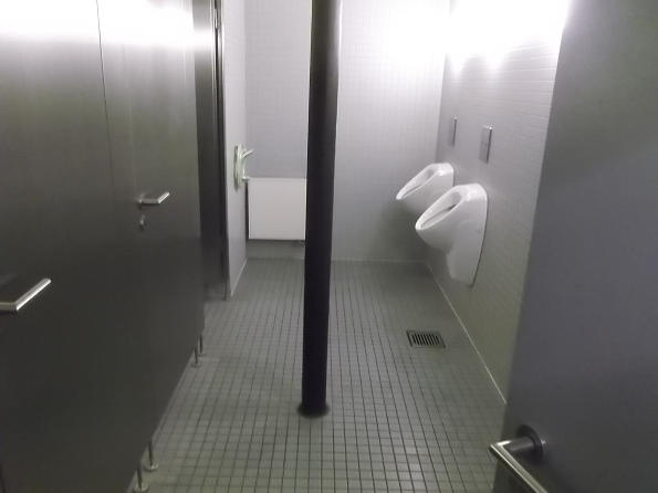 Bild 1 zeigt den Blick in eine Herrentoilette, in dessen Mitte ein schwarzer Mast steht.