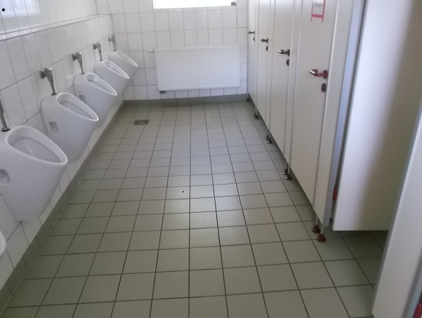 Bild 2 zeigt den Blick in ein Herren WC - links an der Wand hintereinander mehrere Urinale, mittig der geflieste Boden und rechts Türen zu den einzelnen WCs.