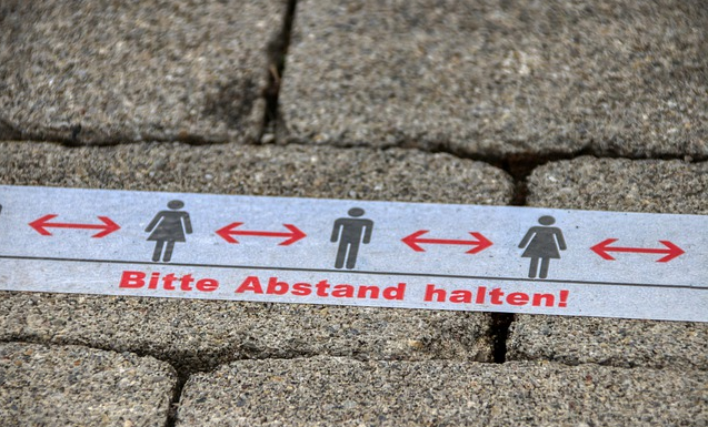 Bild1 zeigt ein weißes Band mit roter Aufschrift - Bitte Abstand halten und darüber rote Pfeile zwischen einzelnen Piktogrammen von Frau und Mann, was auf einen Gehweg aufgeklebt wurde. 