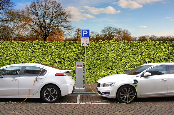 Bild 1 zeigt eine Ladestation auf einem Gehweg vor einer Hecke, wo 2 weiße parkende Autos geladen werden. 
