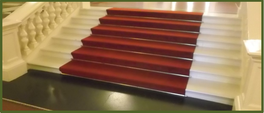 Bild 1 zeigt eine Treppe, auf der in der Mitte auf den Stufen ein roter Teppich liegt. 