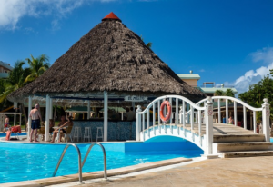 Bild 1: Schwimmbekenumlauf mit einer Treppe zur Gastronomieeinrichtung. Tische und Stühle vor einem Gastronomie-Pavilion mit Reetdach. 