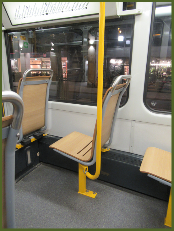 Bild 3 zeigt in einer Straßenbahn eine gelbe vertikale Haltestange und einen Festhaltegriff aus Edelstahl an einer Sitzrückenlehne im visuellen Kontrastvergleich.