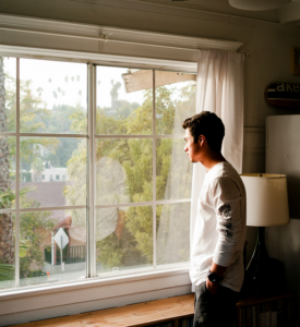 Bild 2 zeigt einen Mann im Wohnzimmer, der auf der rechten Seite eines großen Fensters steht und nach draußen in einen Garten blickt. 