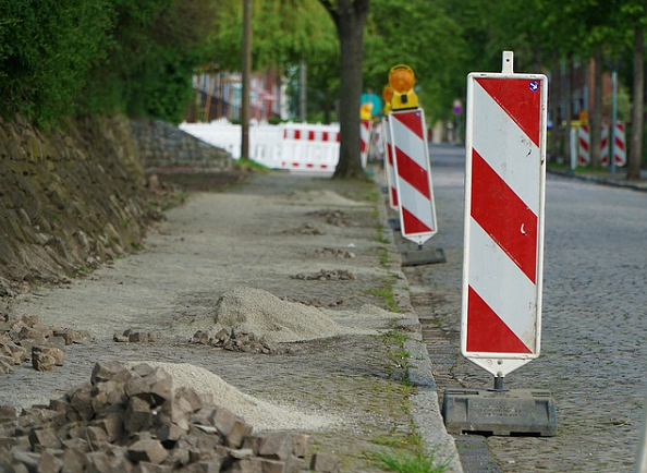 Bild 4: hintereinander stehende rot-weiß gestreifte Schraffenbaken, die den linksseitigen Gehweg abtrennen, auf dem mehrere Steinhaufen liegen 
