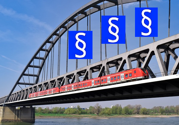 Bild 1: Roter Triebwagen fährt auf Eisenbahnbrücke über einen Fluss, über dem Zug an Stahlseilen blaue Fahnen mit weißem § Zeichen