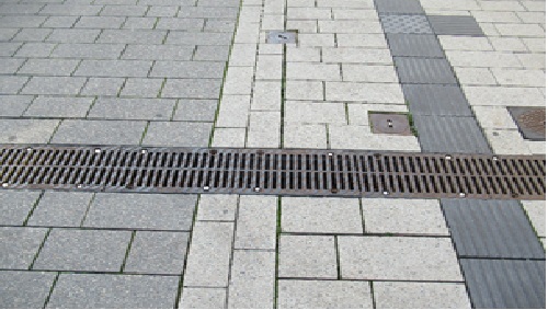 waagerecht verlaufende Entwässerungsrinne mit einer Abdeckung, die Bodenindikatorprofile aufweist, in einer Fußgängerzone, auf der rechten Bildseite horizontal (in Gehrichtung) verlaufender Blindenleitstreifen (Rillen) 